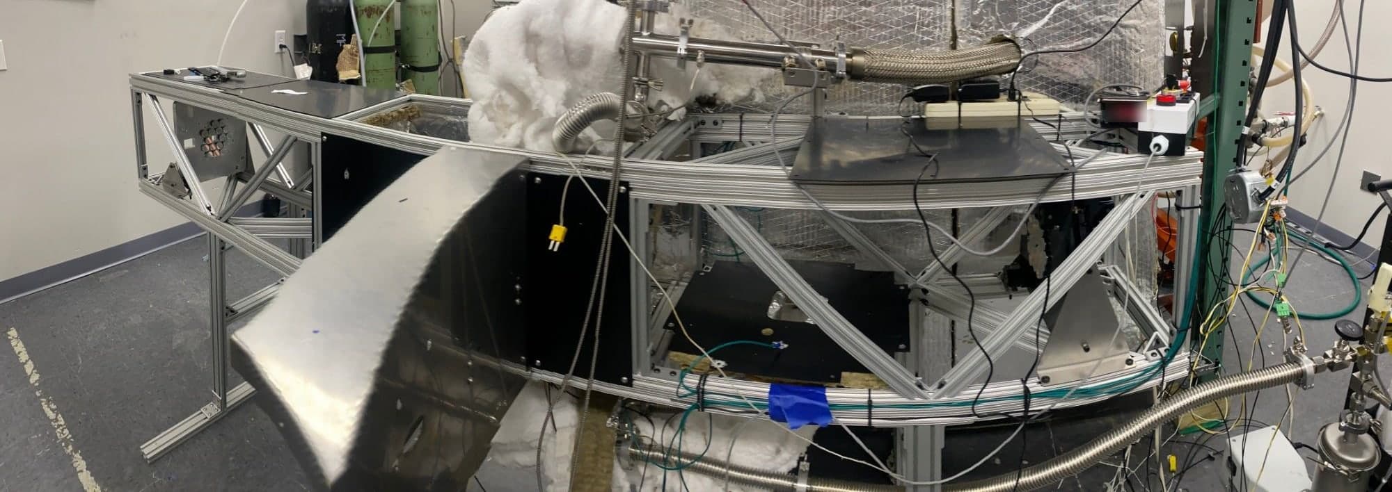 A laboratory apparatus about two meters long, built on an open aluminum frame. A plate holding a cluster of round mirrors stands at its left end. A polished metal housing leans against the front. Behind the right end stands a large vessel wrapped in foil insulation, with white insulation around the gas lines. Gas cylinders stand against the wall.